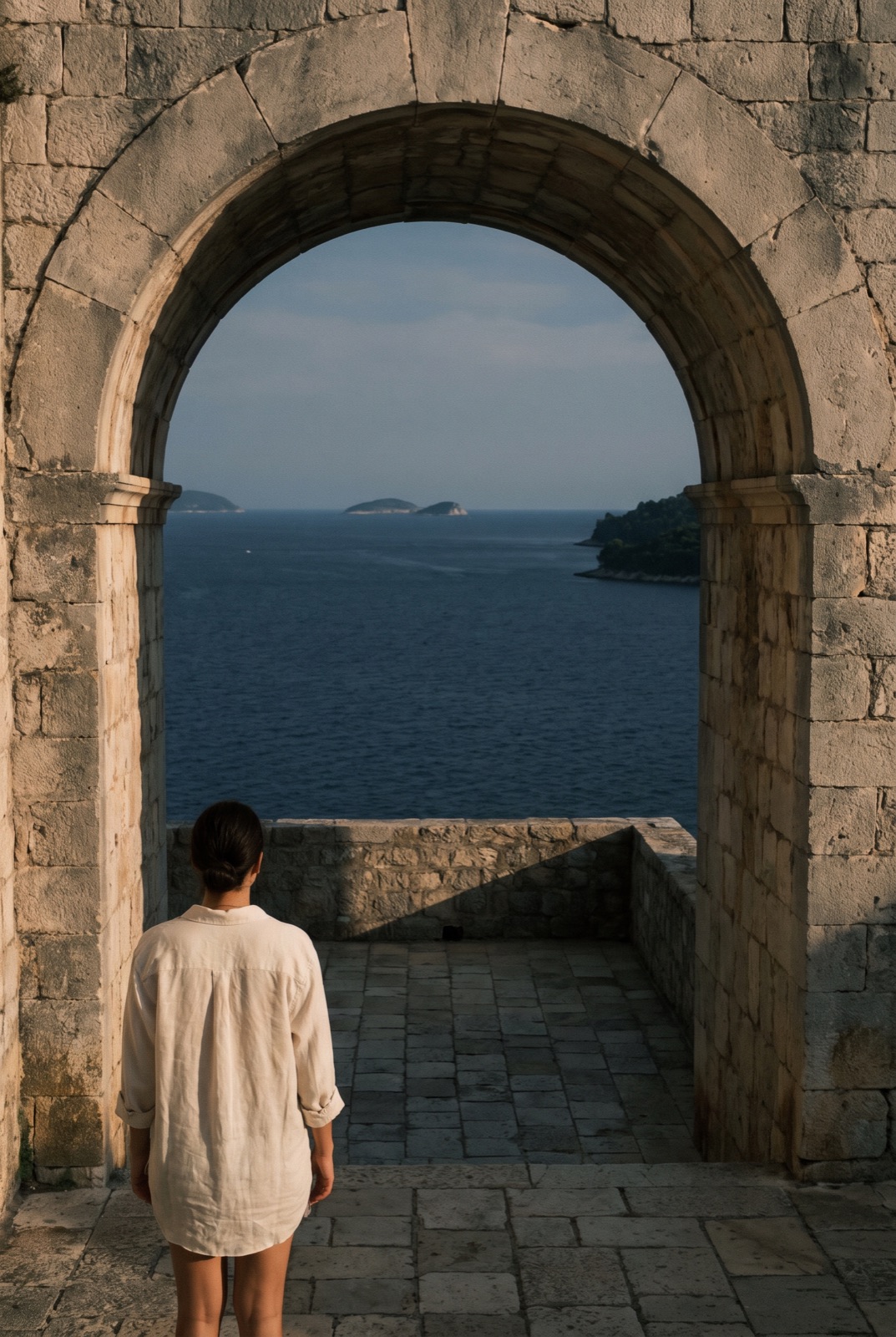 A figure in a stone courtyard, framed by an arch opening onto the Adriatic